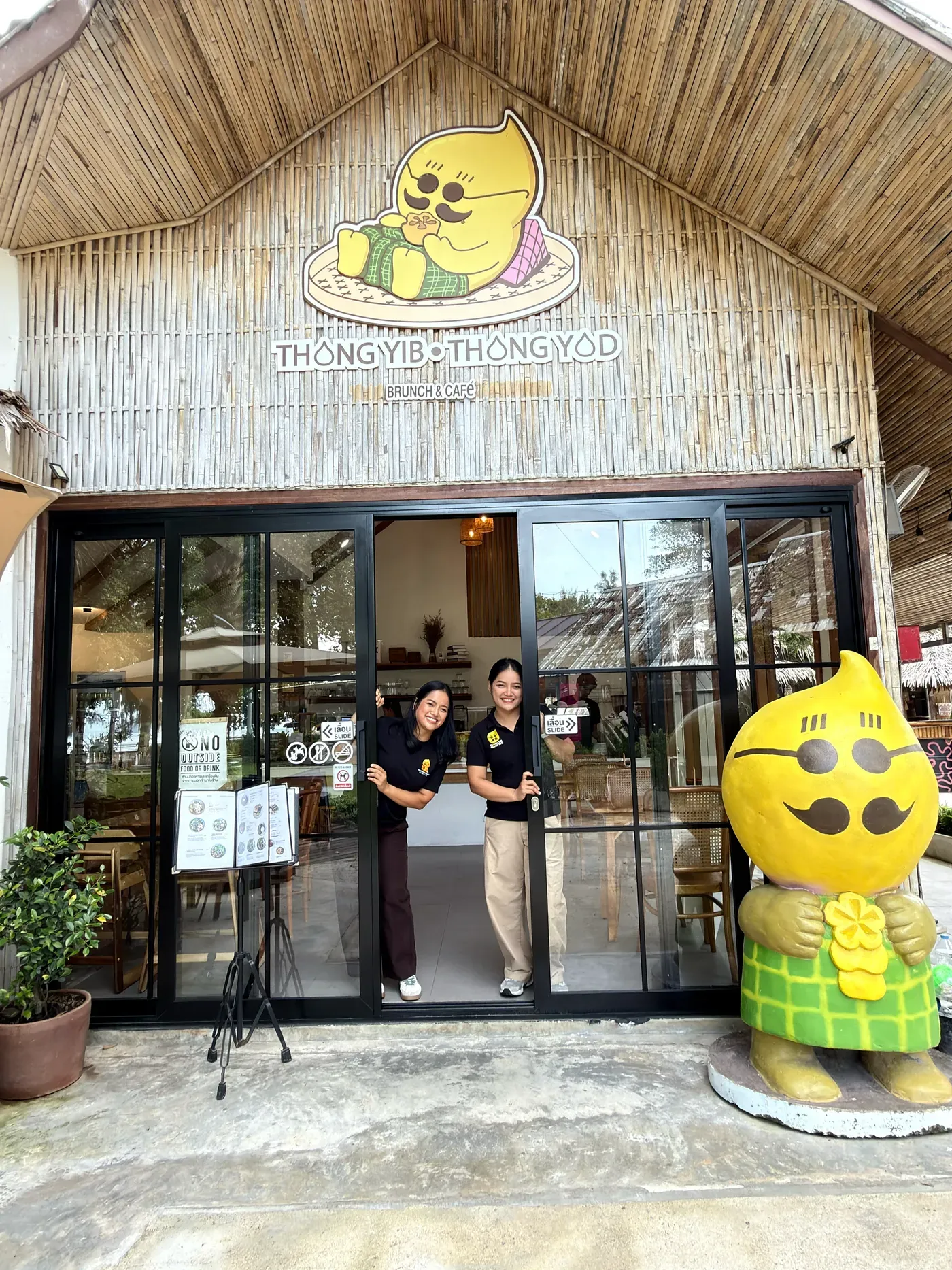 Le sorelle gemelle proprietarie di Thongyib Thongyod sorridono sulla porta del locale accanto alla statua dorata della mascotte