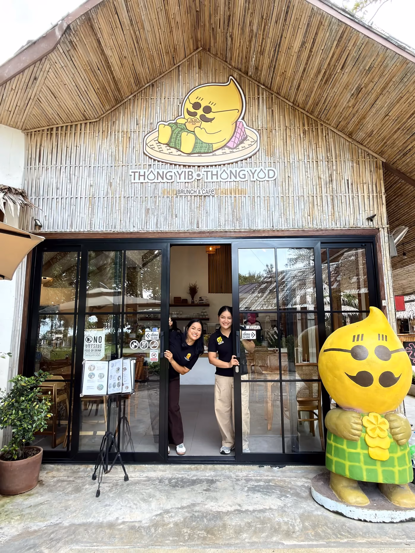 The twin sisters who founded Thongyib Thongyod, smiling and waving from the cafe doorway beside the mascot statue