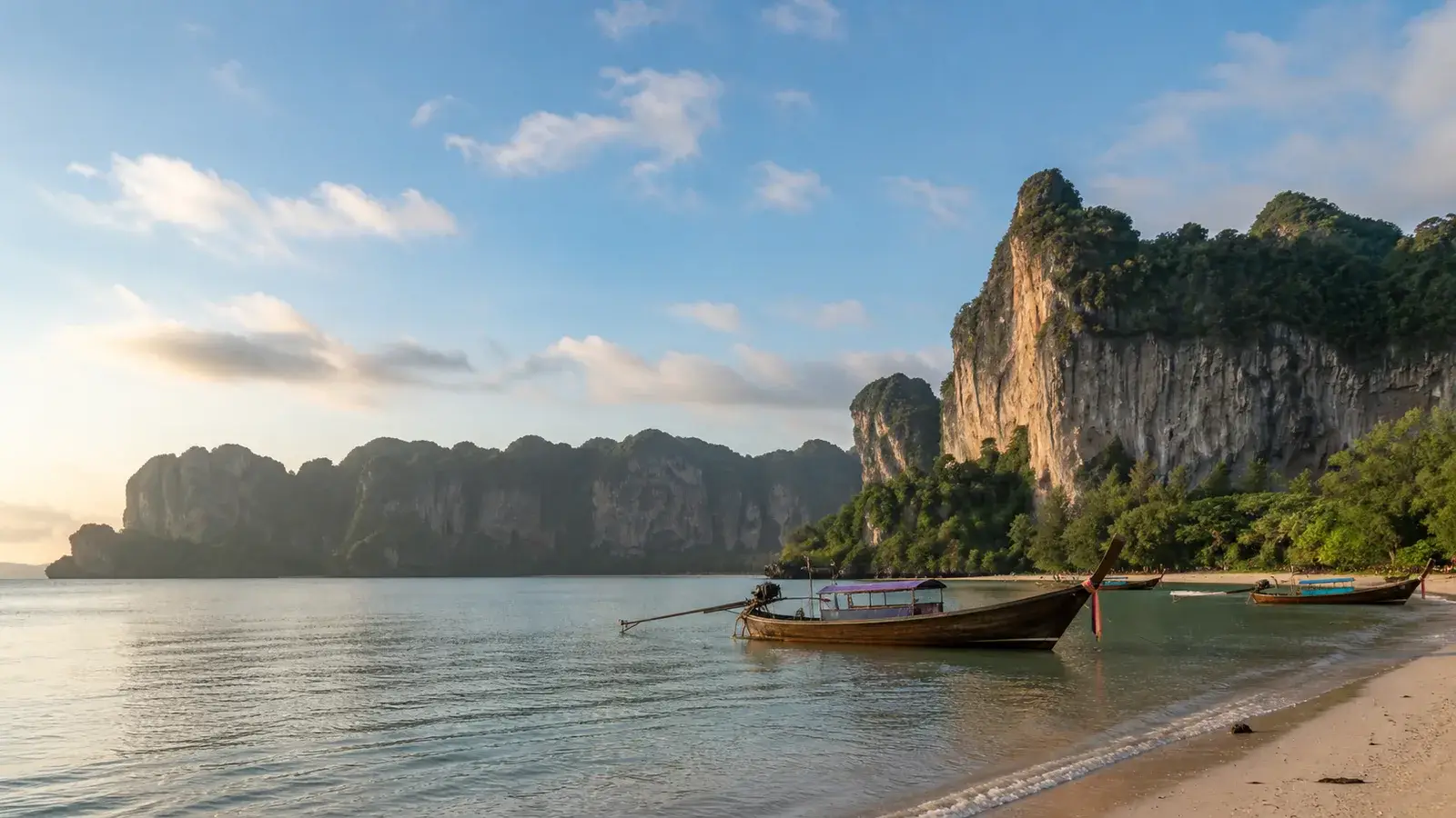 Traditional longtail boats on calm water beneath Krabi limestone cliffs during a clear early dry-season November morning