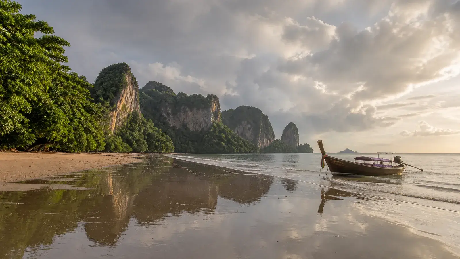 A traditional longtail boat on a reflective Krabi beach after a September rain shower, with green limestone cliffs beneath clearing clouds