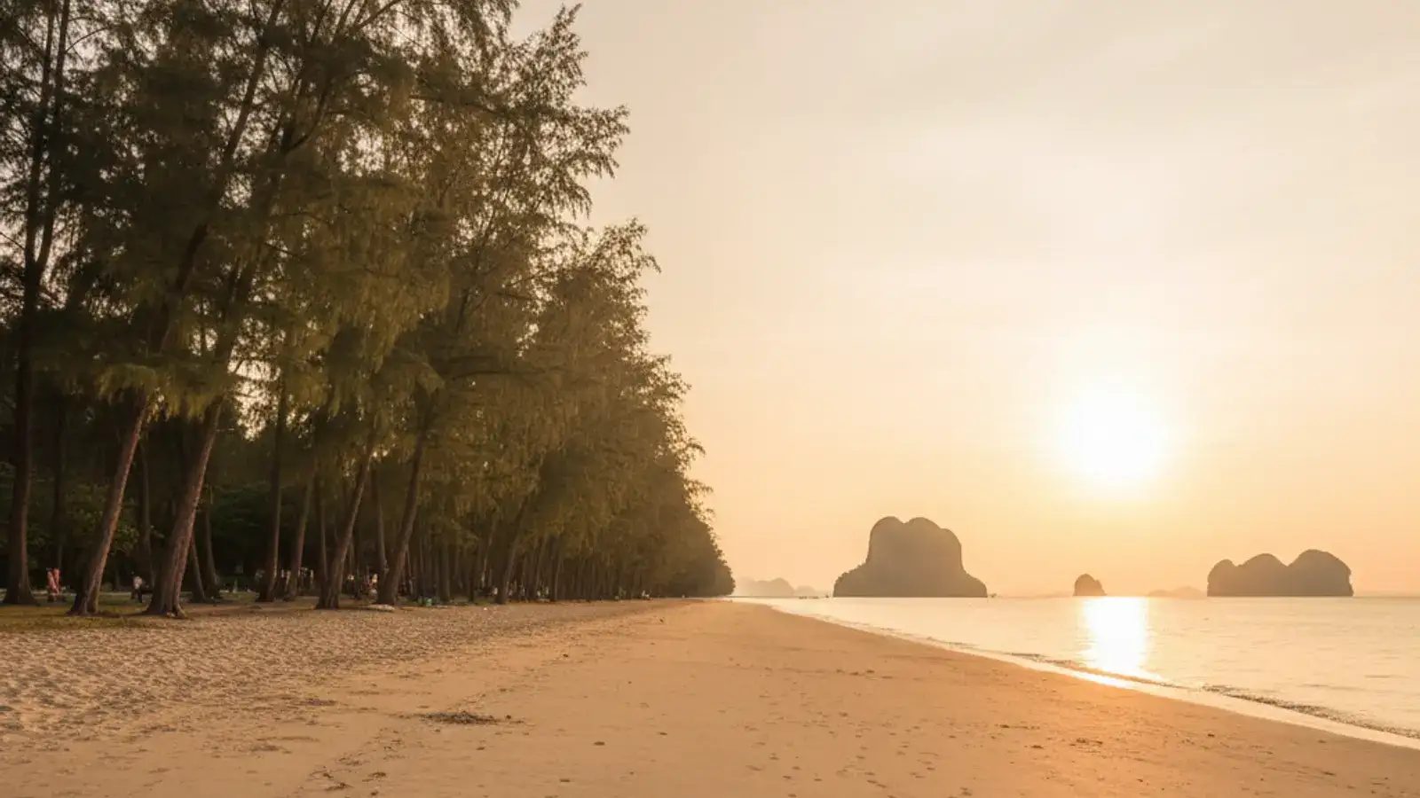 Wide, casuarina-shaded sand at Nopparat Thara Beach at low tide, with a jungle-topped islet offshore and longtail boats in the distance