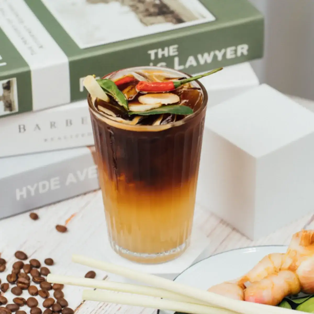 A tall glass of iced tom yum coffee garnished with lemongrass, kaffir lime leaves and a red chili, with coffee beans scattered beside it