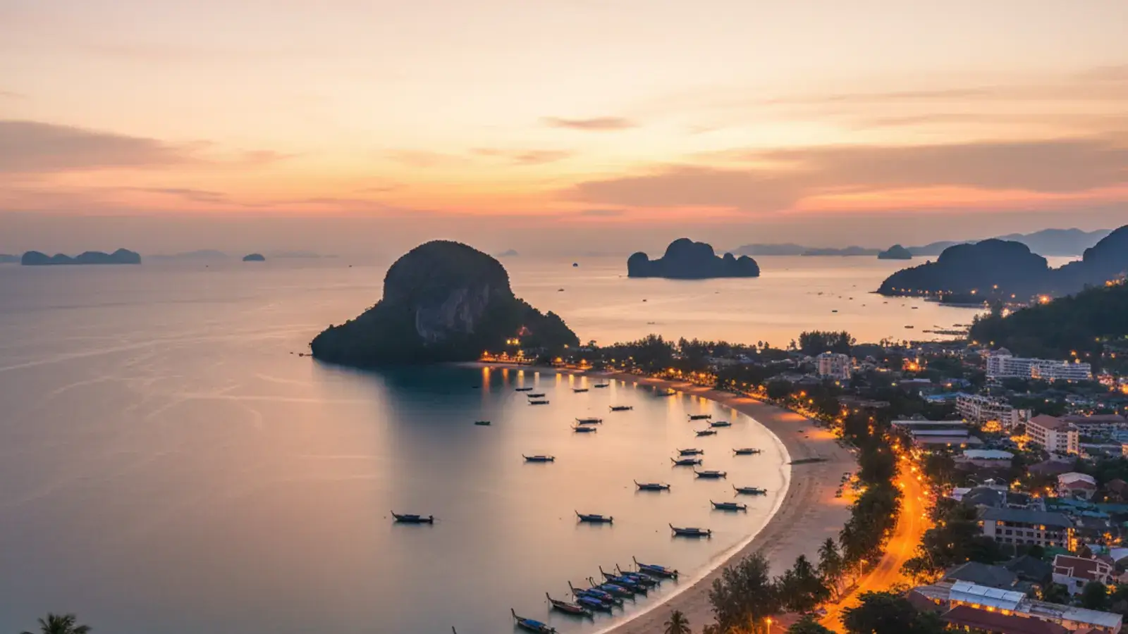 View along the Ao Nang beachfront at golden hour, with longtail boats on the sand and limestone karsts rising behind the bay