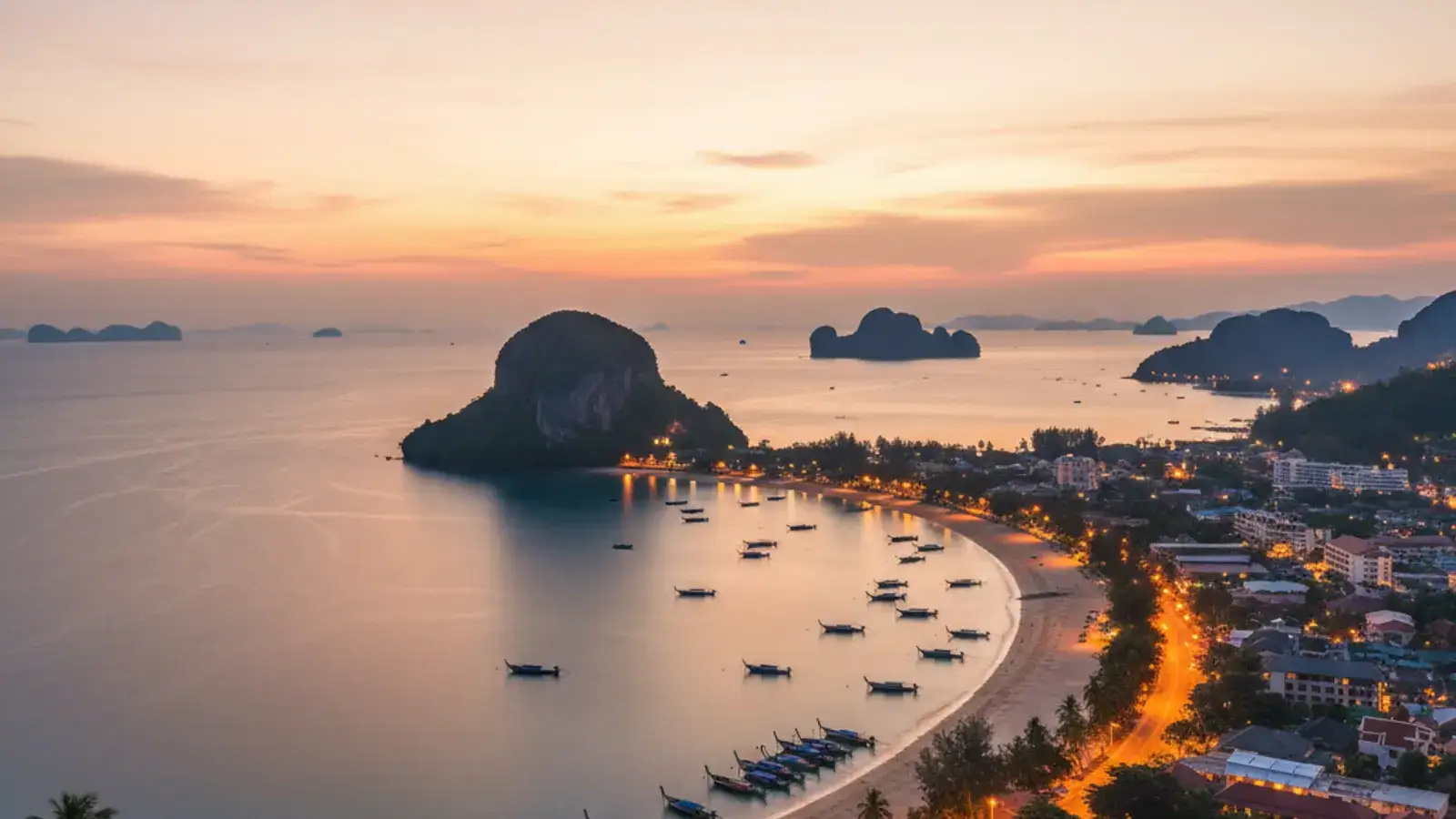 View along the Ao Nang beachfront at golden hour, with longtail boats on the sand and limestone karsts rising behind the bay