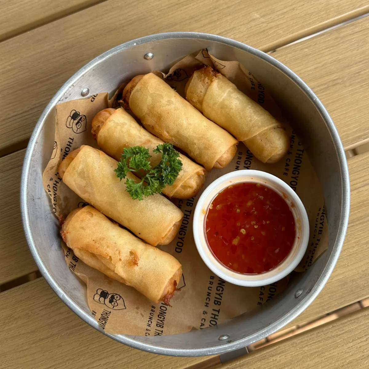 Golden fried spring rolls with a bowl of sweet chilli dip, served in a metal basket lined with branded paper