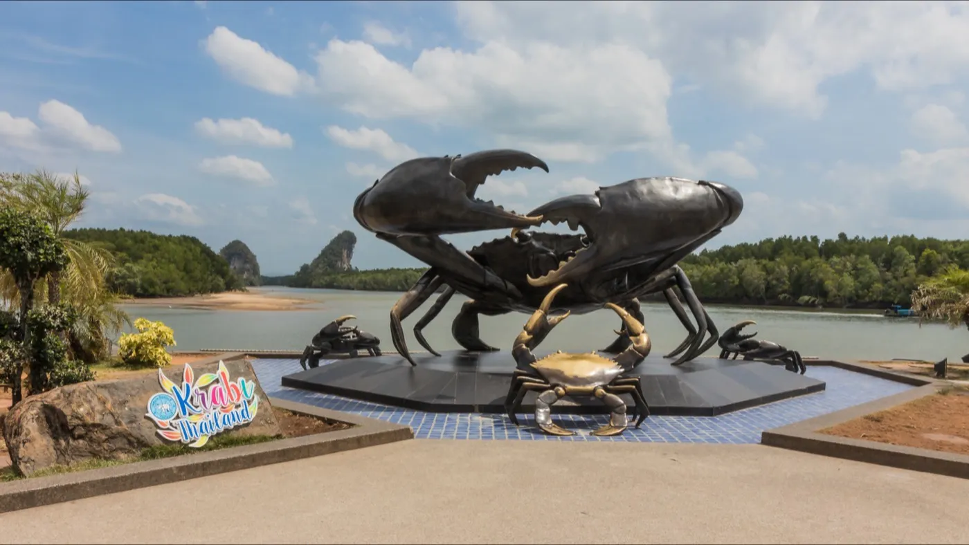 Eine große schwarze Krabben-Skulptur an der Uferpromenade von Krabi, Mangroven und ein Kalksteinhügel auf der anderen Wasserseite dahinter