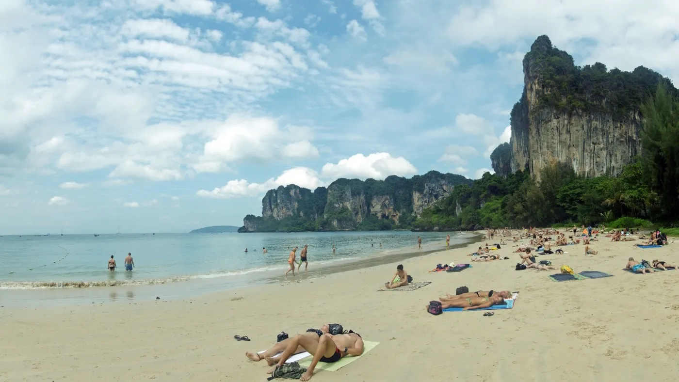 Railay West beach on a bright dry-season day, sunbathers on white sand beneath a towering limestone cliff under a blue sky