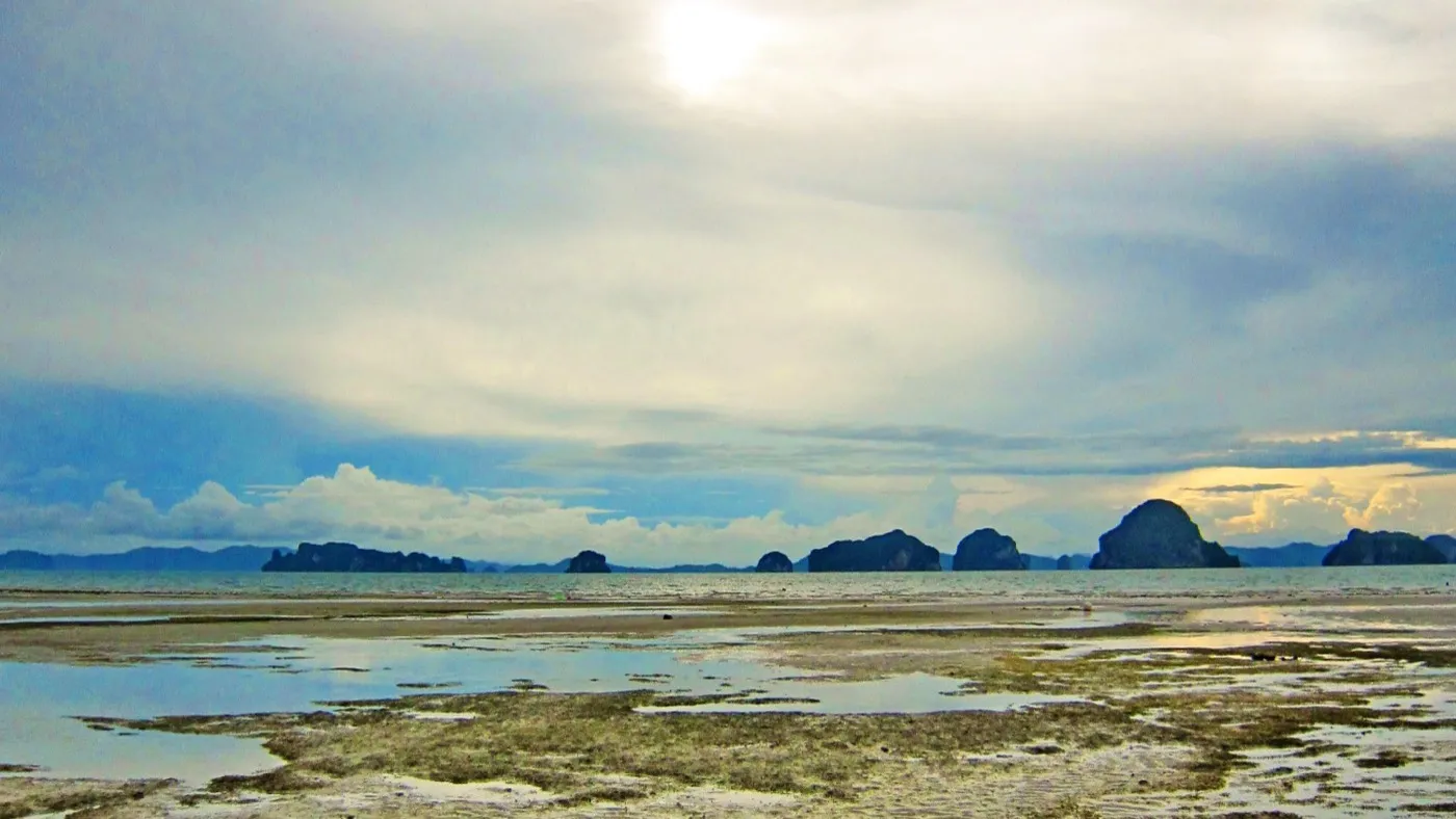 A dramatic monsoon sky over a Krabi beach at low tide, storm clouds massing above silhouetted limestone islands offshore