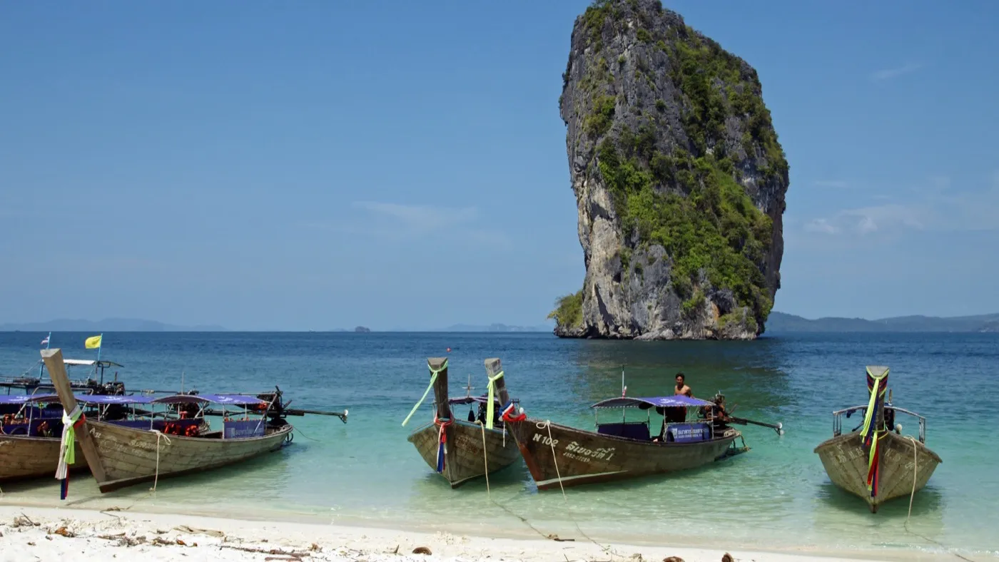 Longtail boats moored off Poda Island's white beach, with the island's tall limestone tower rising from the sea behind them