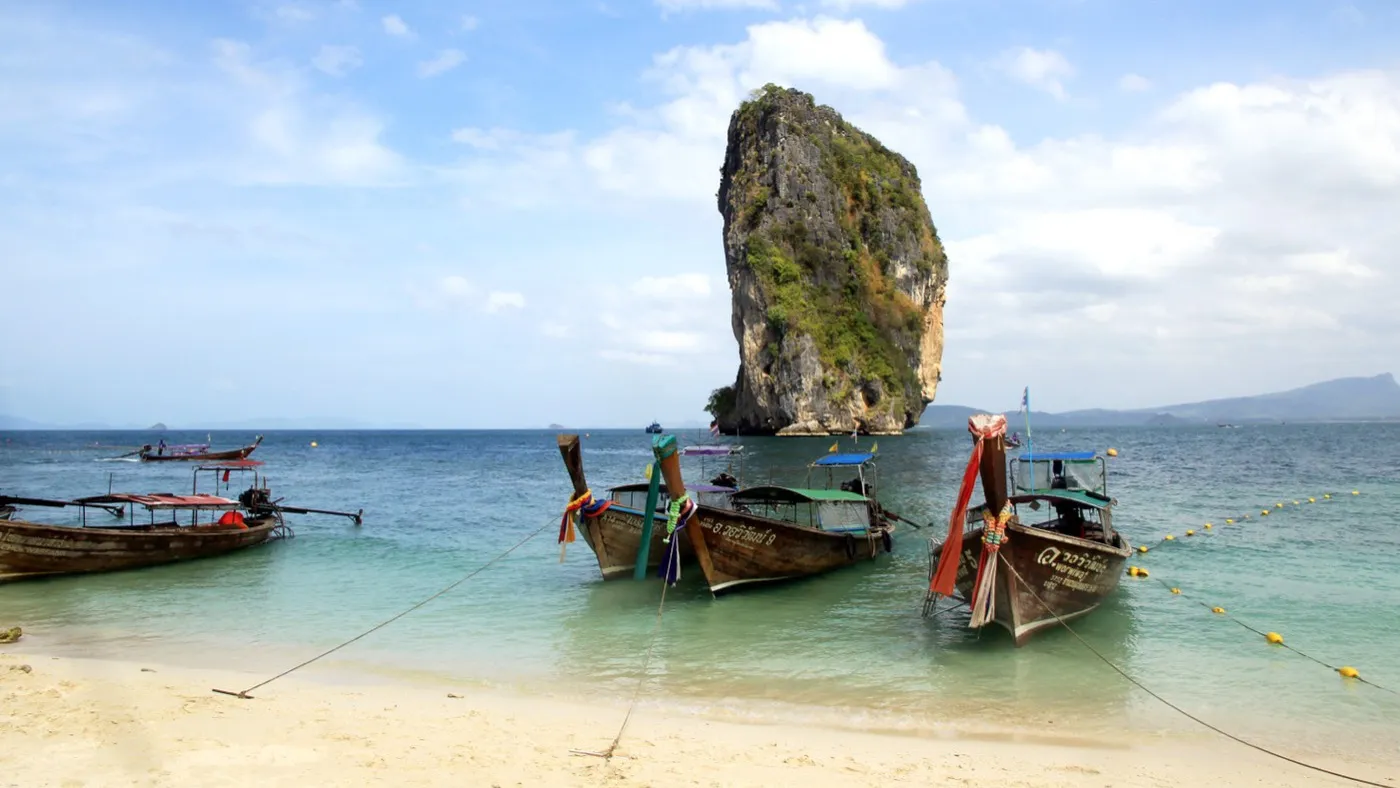 Longtail-Boote vor dem weißen Strand von Poda Island, dahinter ragt ein hoher, dschungelbedeckter Kalksteinturm aus dem türkisfarbenen Wasser