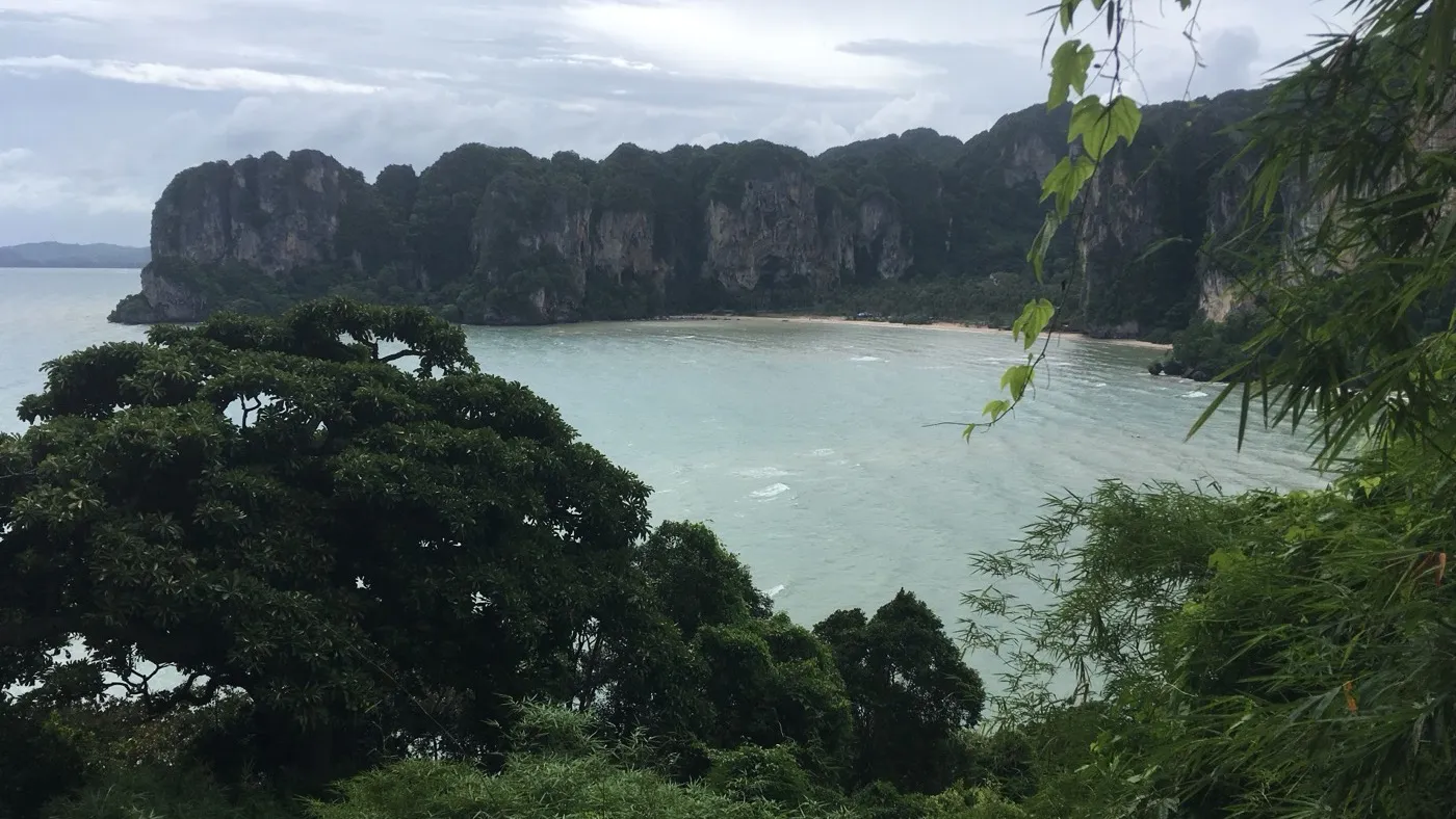 View over Railay bay from a hillside, jungle-topped limestone cliffs curving around a beach with pale green sea below