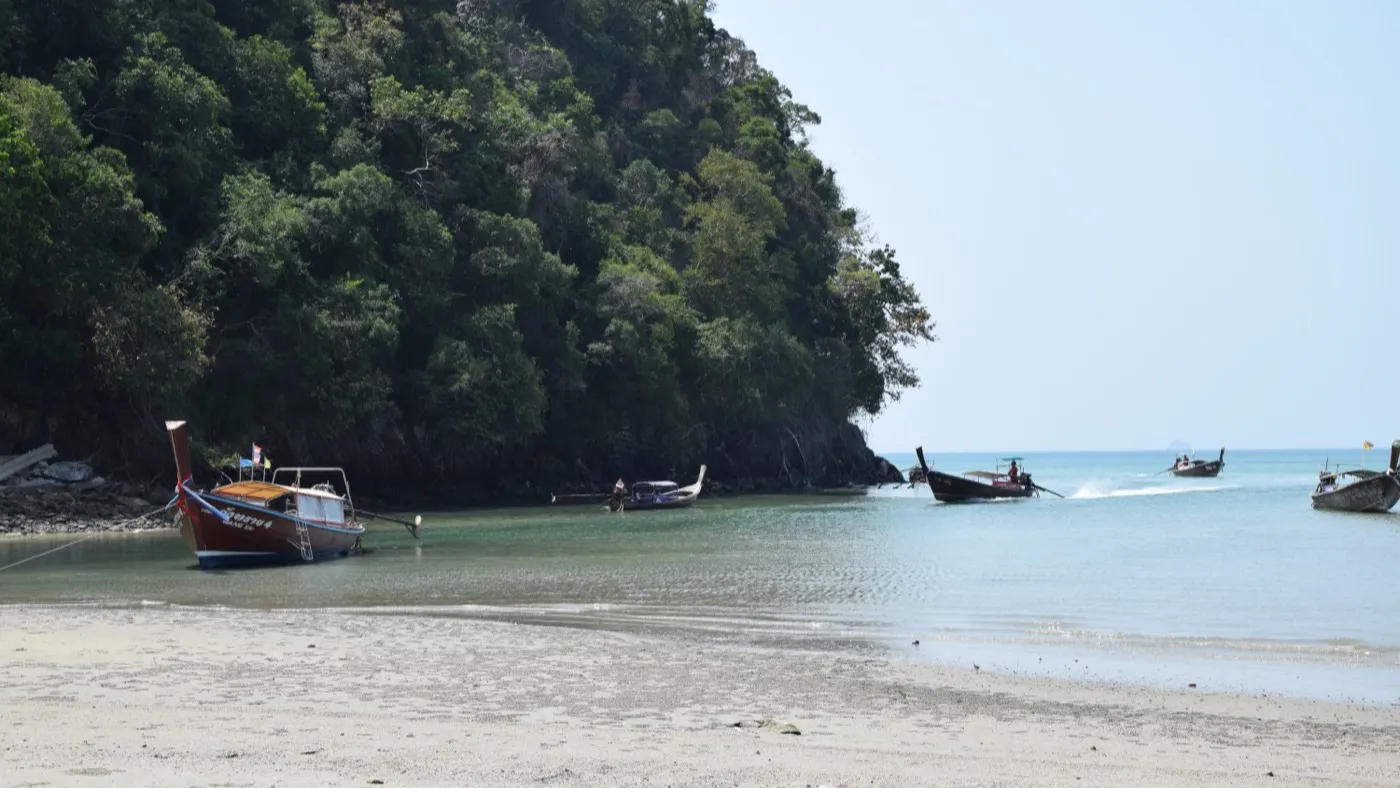 Longtail-Boote ruhen im Sand einer stillen Bucht bei Ao Nang, dahinter erhebt sich eine bewaldete Kalksteinlandzunge