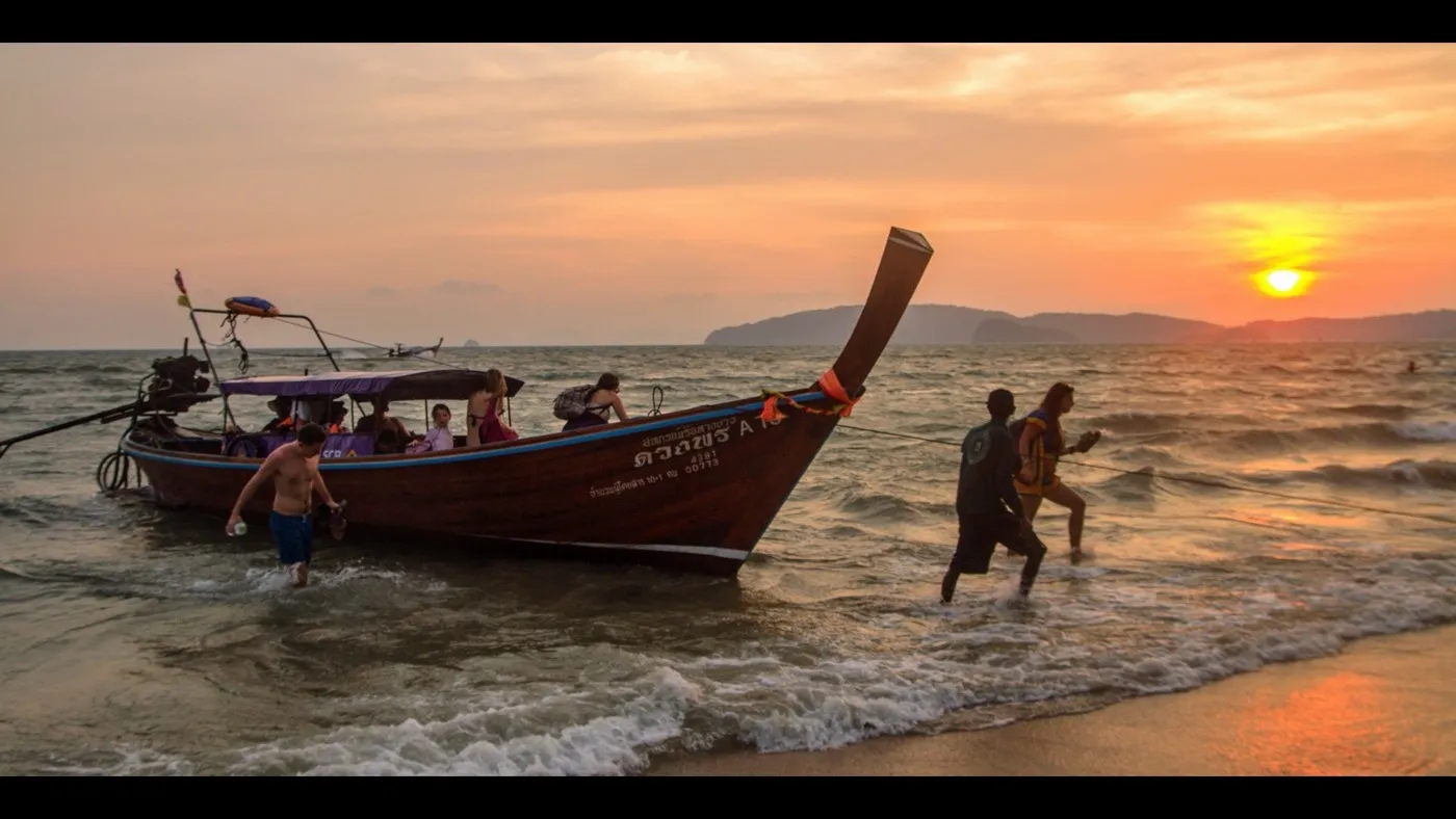Łódź longtail wyciągnięta na plażę Ao Nang o zachodzie słońca, z pomarańczowym niebem nad zarysowanymi wzgórzami i spokojnym morzem