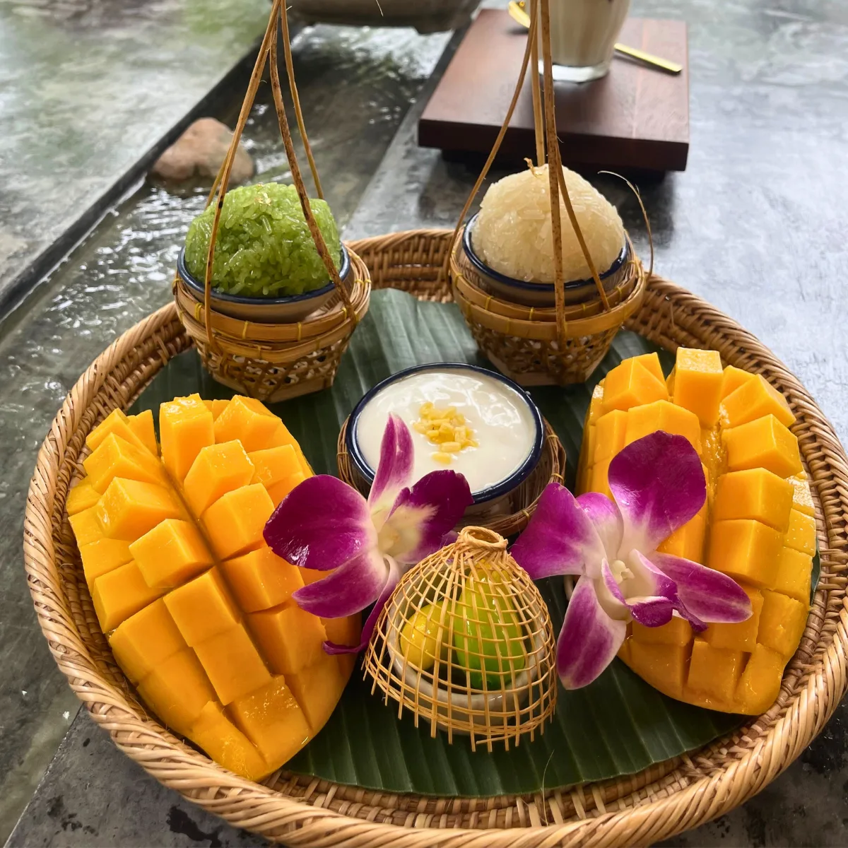 A sharing platter of mango sticky rice with two whole hedgehog-cut mangoes, small baskets of white and pandan-green sticky rice, and a bowl of salted coconut cream, garnished with purple orchids