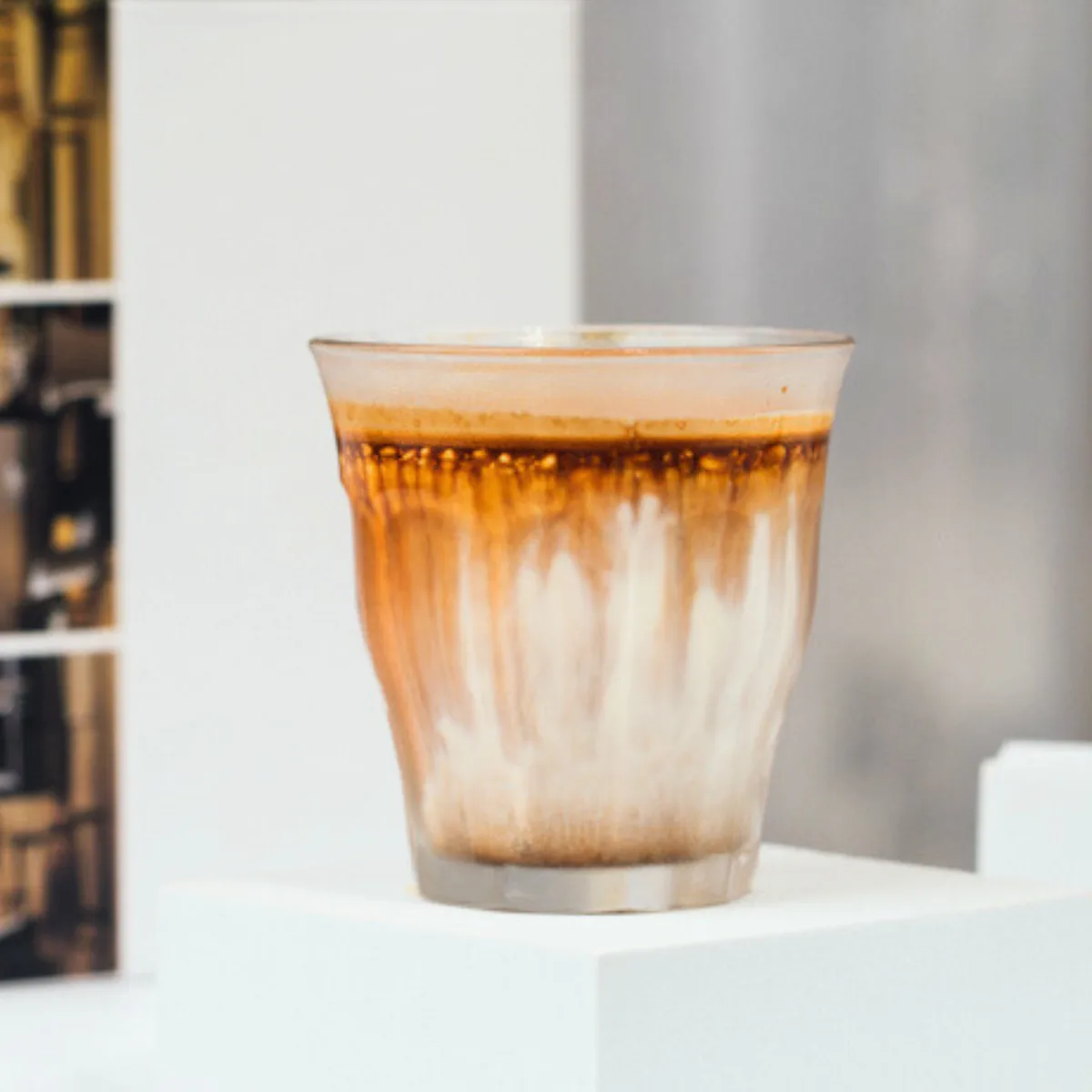 Ein beschlagenes Glas Dirty Coffee — ein dunkler Espresso-Shot, über kalte Milch gegossen, der braune Spuren durch das Weiß zieht — auf einem weißen Podest