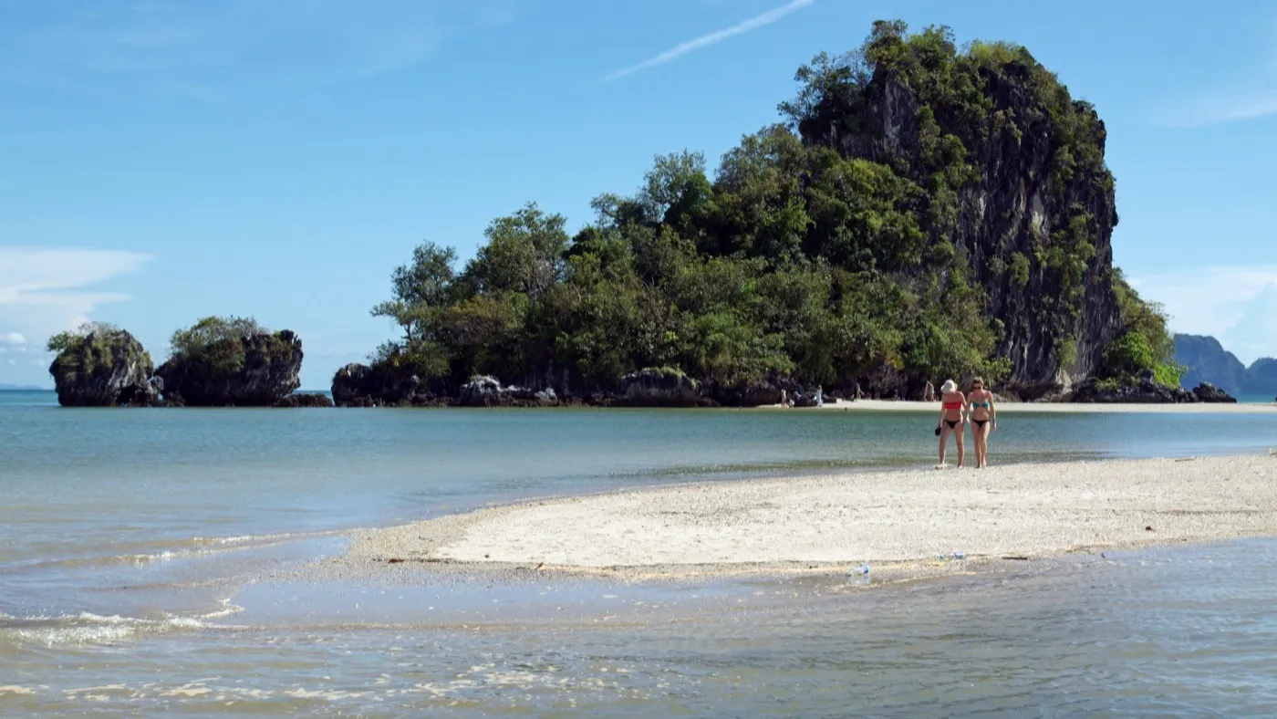 Two people walking across an exposed low-tide sandbar towards a jungle-topped limestone islet off Nopparat Thara Beach