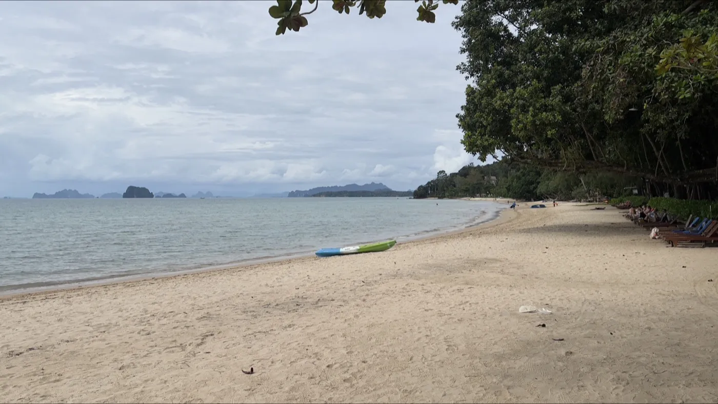 Den lugna sanden och det stilla vattnet vid Tubkaek Beach, inramat av träd, med kalkstensöar på andra sidan bukten
