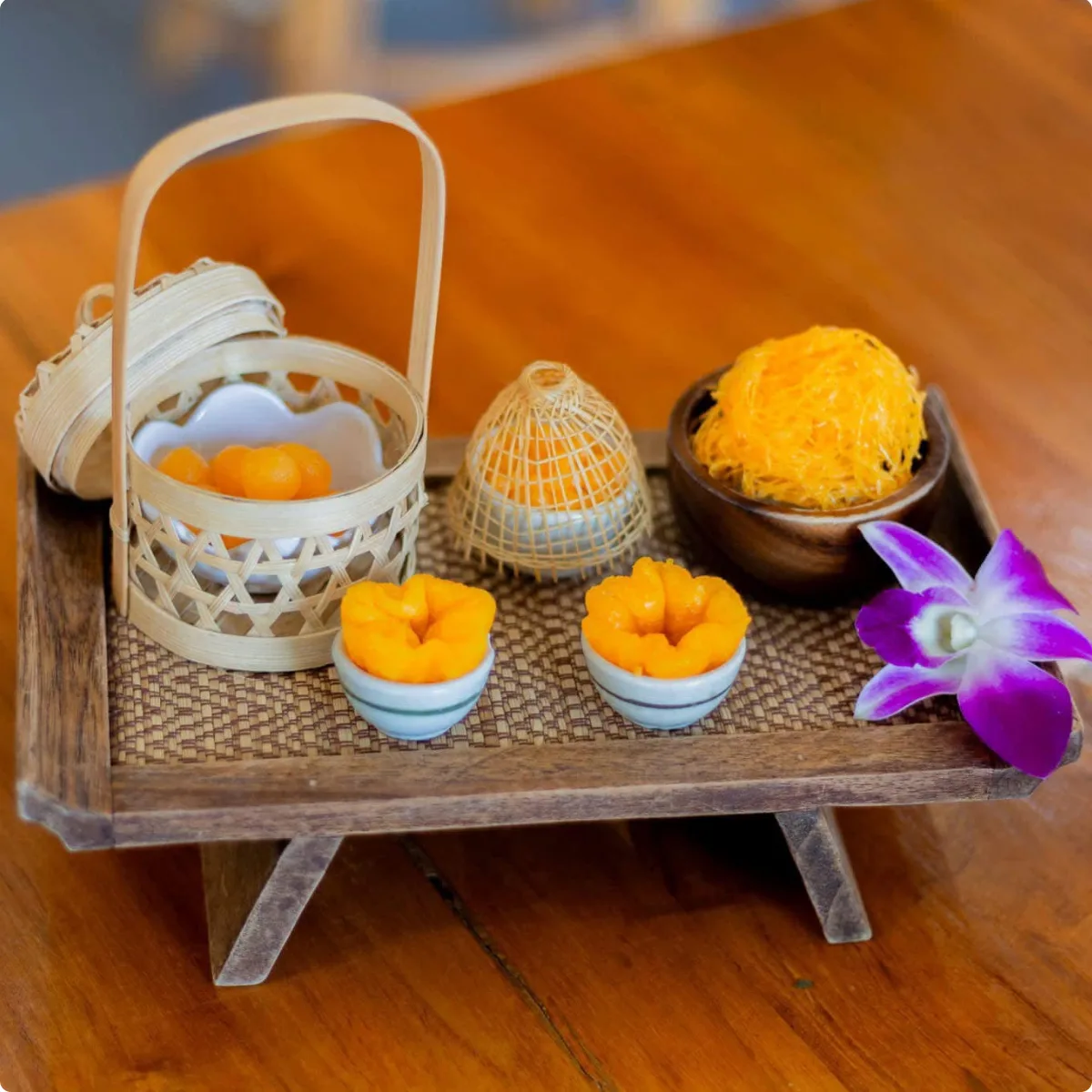 A wooden tray of golden Thai egg-yolk desserts — glossy thong yod teardrops in a basket, pinched thong yip flowers in tiny cups, and a nest of fine foi thong threads — with a purple orchid