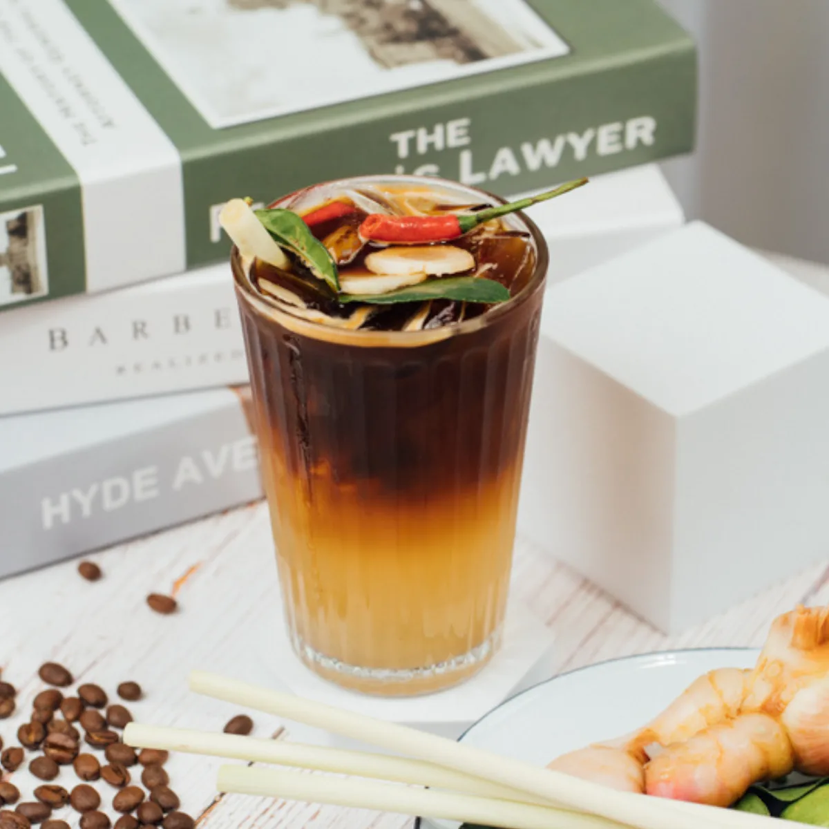 Ein hohes Glas eisgekühlter schwarzer Kaffee, garniert mit Zitronengras, Kaffirlimettenblättern und einer roten Chili, daneben verstreute Kaffeebohnen und Garnelen