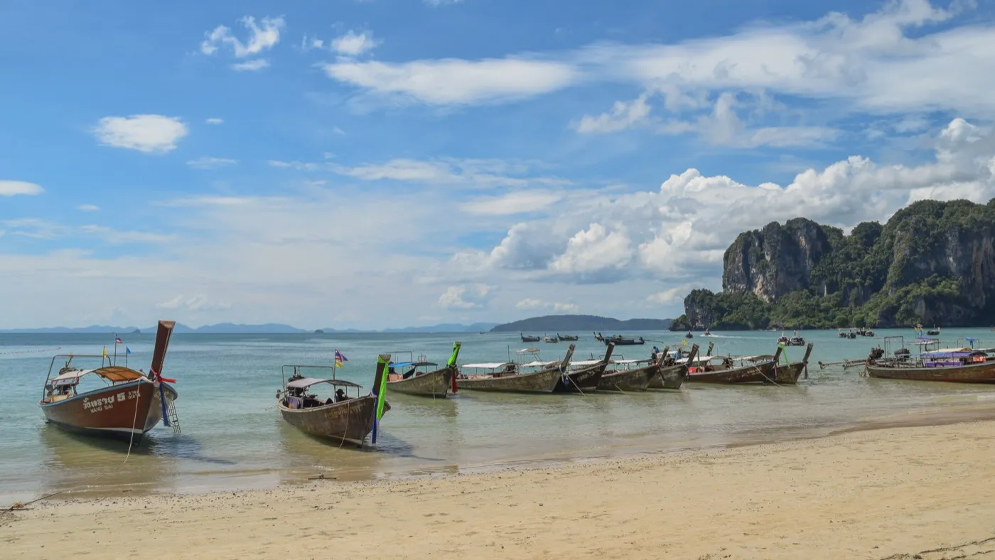 Longtail boats lined up on the sand at Railay West beneath a jungle-topped limestone cliff
